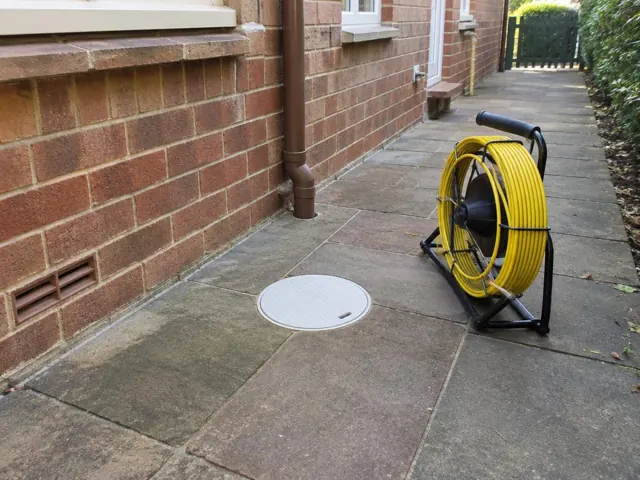 Capped inspection opening on a paved side path beside a coiled drain camera reel and brick wall in Normanhurst