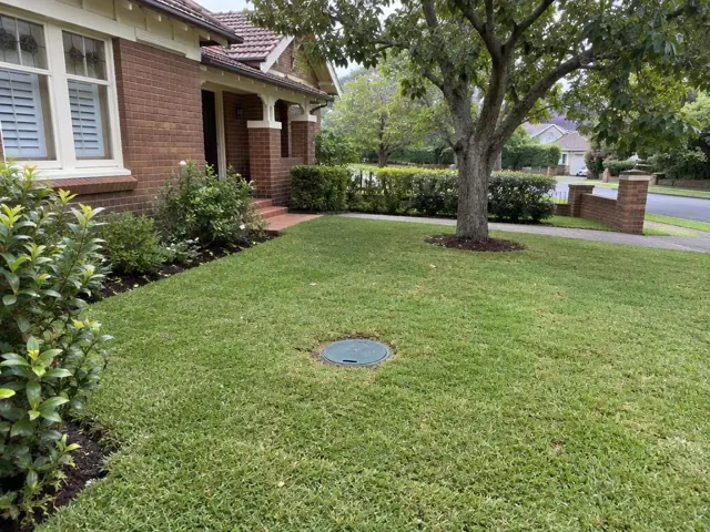 Capped sewer access cover in a neat front lawn beside a brick house in Normanhurst